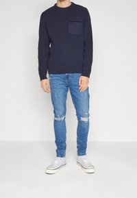 Jersey de punto acanalado en color azul marino con un bolsillo en el pecho, combinado con unos vaqueros desgastados de color azul claro y zapatillas blancas con detalles en negro.