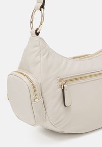 Sac à épaule beige avec des fermetures éclair dorées, une petite poche avant et un anneau en métal doré reliant la bandoulière au corps du sac.