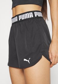 Close-up van een persoon die zwarte Puma-shorts draagt met een merkbare tailleband en een wit Puma-logo bij de zoom, hand rustend aan de zijkant.