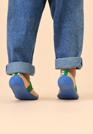 Grüne Sandale mit atmungsaktiven Riemen, Korkfußbett und blauer Gummisohle. Hellbraune Akzente an den Seiten. Hochgekrempelte Jeans sichtbar darüber.