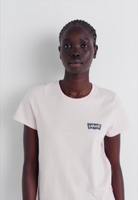 T-shirt en coton rose clair à manches courtes, avec un patch logo Levis bleu sur la zone gauche de la poitrine. Texture lisse, coupe standard.