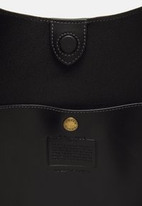 Interior de bolso de cuero negro con cierre de botón a presión cosido, botón dorado con el logo de Coach y etiqueta Coach en relieve con número de serie.