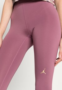 Träningsleggings i en dämpad lila färg, med en gul Jordan-logotyp, slät textur och kontrasterande sömmar längs sömmarna.
