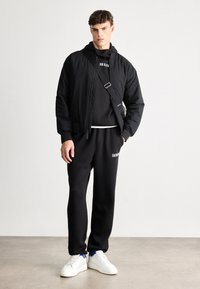 Czarna bomberka z zapięciem na zamek, w połączeniu z czarną bluzą z kapturem oraz dresami z białym napisem, dopełnione białymi sneakersami.