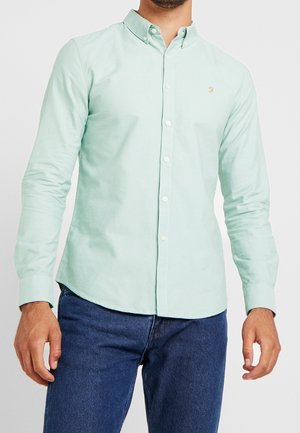 Homme portant une chemise vert clair boutonnée à manches longues et un jean bleu foncé sur un fond uni.