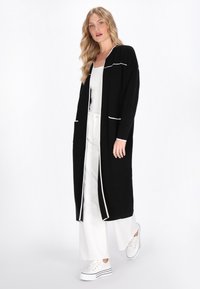 Schwarzer langer Cardigan mit weißem Saum, offene Vorderseite, zwei Taschen, über einem weißen Oberteil und weiten weißen Hosen getragen. Modell trägt weiße Sneaker.