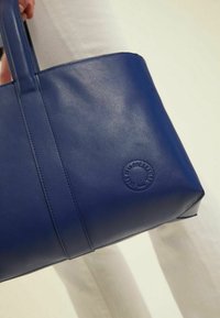 Sac fourre-tout en cuir bleu avec deux poignées, présentant un logo embossé arrondi, une texture lisse et une forme angulaire.