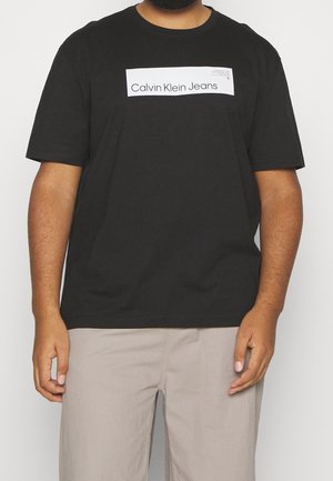 Homme portant un t-shirt noir à manches courtes avec le logo blanc "Calvin Klein Jeans" sur la poitrine et un pantalon beige clair, sur un fond uni.