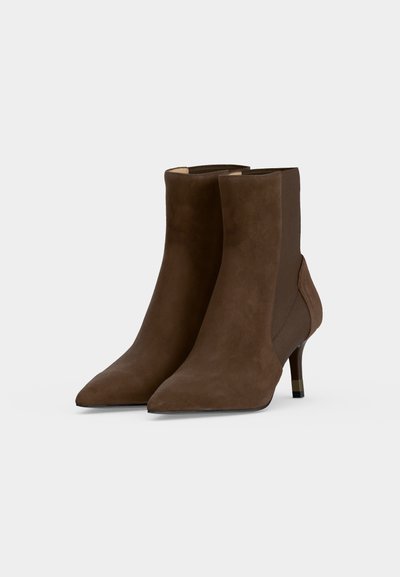 Paire de bottines en daim marron à bouts pointus, talons fins de hauteur moyenne et panneaux latéraux élastiques sur un fond clair uni.