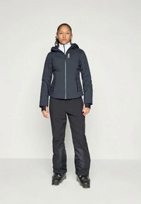 Schwarze isolierte Skijacke mit Reißverschluss, Kapuze und Logodetail, kombiniert mit passenden dunklen Hosen und schwarzen Skischuhen. Flache Textur, enganliegendes Design.