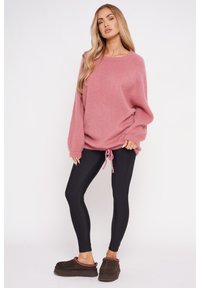 Jasnoróżowy, oversize sweter z długimi rękawami i ściągaczem na dole, w połączeniu z czarnymi legginsami i brązowymi butami wsuwanymi.