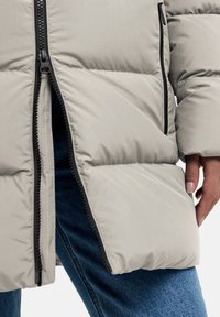 Lichtgrijze pufferjack met zwarte ritsdetails, met een gestikt ontwerp en een close-up van een hand die de zoom vasthoudt. Gecombineerd met blauwe jeans.