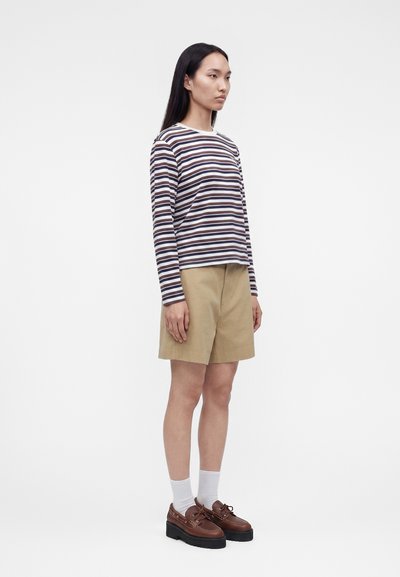 Γυναίκα που στέκεται πλάγια, φοράει μια μπλούζα με καφέ, λευκές και ναυτικές ρίγες με μακριά μανίκια, μπεζ σορτς, λευκές κάλτσες και καφέ παπούτσια με πλατφόρμα.