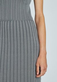 Graues geripptes Kleid mit einem figurbetonten Oberteil und einem schwingenden Rock. Strukturierten Stoff mit vertikalen Streifen. Hand ruht an der Seite.