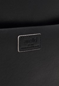 Svart texturerad konstläderyta med en sömd rektangulär etikett som visar varumärket "anello" på en väska eller accessoar.