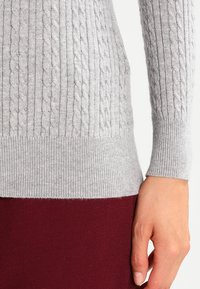 Maglione lavorato a maglia grigio chiaro con motivo a treccia sulle maniche, polsi a costine e orlo. Abbinato a una gonna bordeaux.