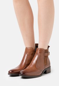 Hnědé kožené kotníkové boty s elastickými panely na stranách a ozdobným páskem, s nízkým blokovým podpatkem a hladkým povrchem.