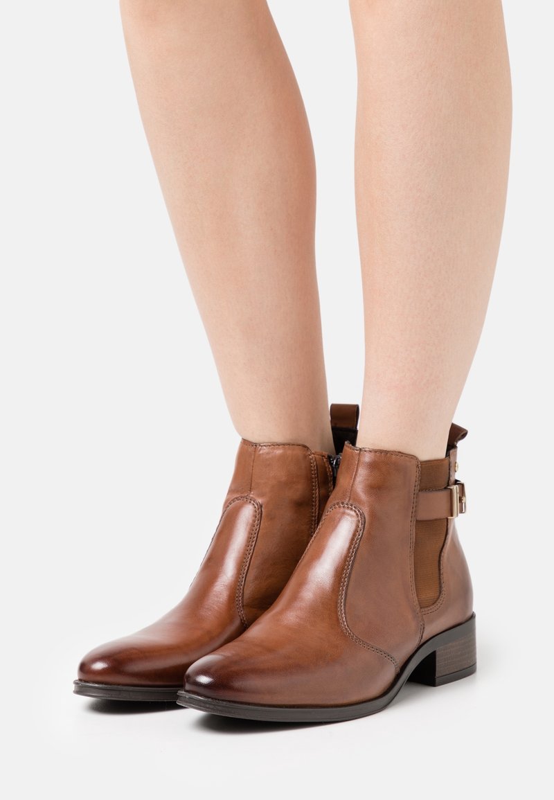 Hnědé kožené kotníkové boty s elastickými panely na stranách a ozdobným páskem, s nízkým blokovým podpatkem a hladkým povrchem.