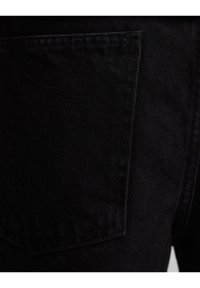 Jeans de mezclilla negra con un bolsillo trasero, costuras visibles y una superficie de tela texturada. Primer plano de los detalles del material y el diseño.