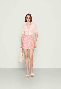 Top rosa claro com mangas bufantes e fecho de botão, combinado com uma saia mini floral em tons de rosa e laranja. A modelo segura uma mala bege.