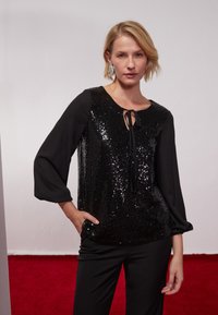 Schwarze Bluse mit einem glänzenden Vorderteil, rundem Halsausschnitt mit Schleifenverschluss und langen Satinärmeln. Kombiniert mit enganliegenden schwarzen Hosen.