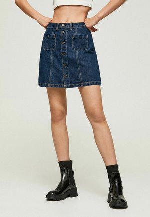 Vrouw die een denim minirok met knopen aan de voorkant en grote zakken draagt, zwarte enkellaarzen en een kort wit topje met de handen in de zij.