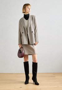 Blazer à carreaux avec boutons doubles, superposé à un pull léger, associé à une jupe beige, des bottes noires montantes et un sac bordeaux.