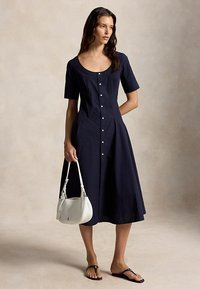 Γυναίκα με μεσάτο φόρεμα μπλε ναυτικού χρώματος με κουμπιά μπροστά, κρατώντας λευκή τσάντα χειρός, φοράει μαύρα σανδάλια, στέκεται μπροστά σε ουδέτερο φόντο.