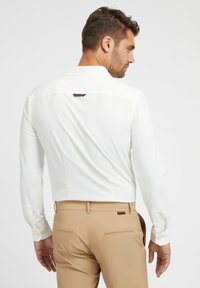 Camicia bianca a maniche lunghe con polsini a bottoni, caratterizzata da un logo sottile sul retro. Abbinata a pantaloni beige e una texture liscia.