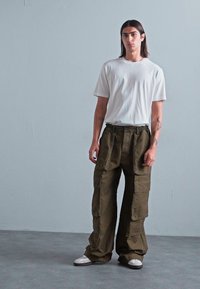 Junge Person mit langen dunklen Haaren steht gegen eine graue Wand und trägt ein weißes T-Shirt, olivgrüne Cargo-Hosen und weiße Sneaker.