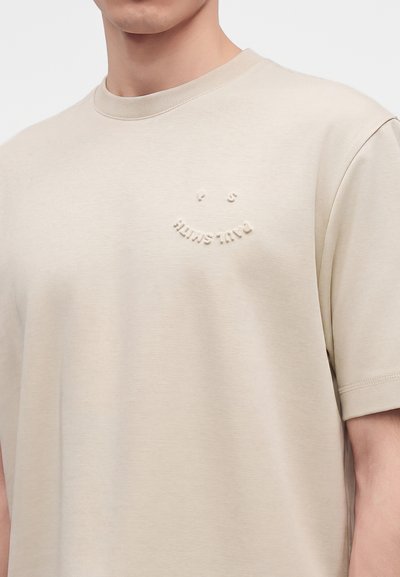 Μπεζ κοντομάνικο t-shirt με μικρό, ανάγλυφο χαμογελαστό πρόσωπο και κείμενο στο αριστερό στήθος, που φοριέται από ένα άτομο από τον λαιμό μέχρι τη μέση του κορμού.