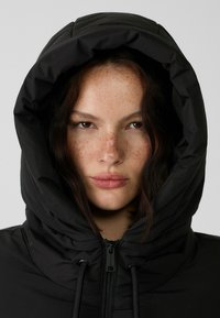 Doudoune noire avec une grande capuche, dotée d'une fermeture éclair à l'avant et de cordons de serrage pour un ajustement personnalisable ; texture lisse et design minimal.
