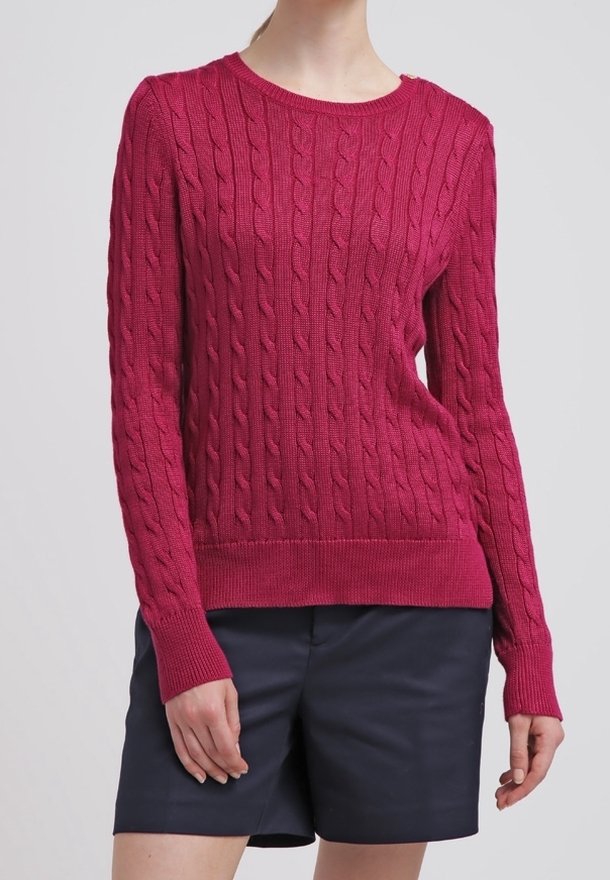 Femme portant un pull rouge à manches longues en maille torsadée associé à un short noir, debout devant un fond gris uni.