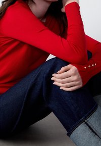 Red knitted sweater with ribbed cuffs featuring gold button accents, paired with dark denim jeans, sitting pose on a gray surface.