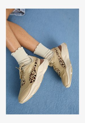 Nohy překřížené, obuté v béžových teniskách Puma s leopardím vzorem pruhů a krémových ponožkách s volánky na modrém koberci.