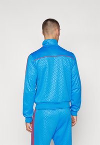 Modrá sportovní bunda s vysokým límcem, texturovaným vzorem a červenými pruhy podél ramen a rukávů. K tomu odpovídající modré kalhoty.