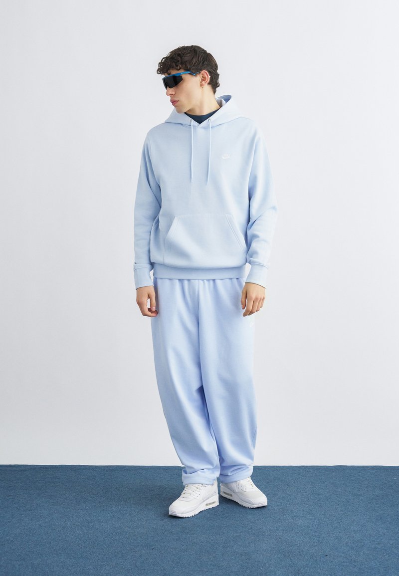 Joven con sudadera con capucha azul claro y pantalones deportivos a juego, zapatillas blancas y gafas de sol negras, de pie sobre una alfombra azul contra una pared blanca.