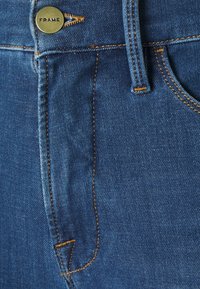 Vaqueros de mezclilla azul con un botón de metal dorado etiquetado como "FRAME", costuras amarillas y diseño clásico de cinco bolsillos con textura visible.