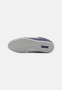 Semelle d'une basket bleu foncé avec une midsole blanche, une semelle extérieure en caoutchouc gris texturé comportant un patch logo Lacoste.