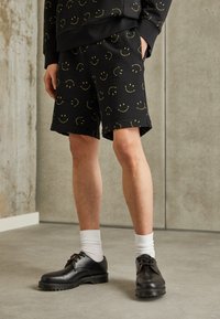 Shorts noirs avec des motifs de visages souriants jaunes ; fabriqués en tissu doux. Associés à des chaussures noires et des chaussettes blanches montantes.