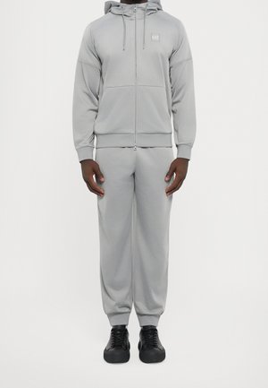 Homme portant un sweat à capuche gris clair zippé et un pantalon de jogging assorti avec des baskets noires, debout sur un fond blanc.