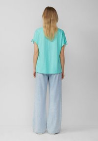 Türkises, lockeres Oberteil mit kurzen Ärmeln und gebundenen Akzenten. Kombiniert mit weiten, hellblauen Denim-Jeans. Minimalistisches Design, glatte Textur.