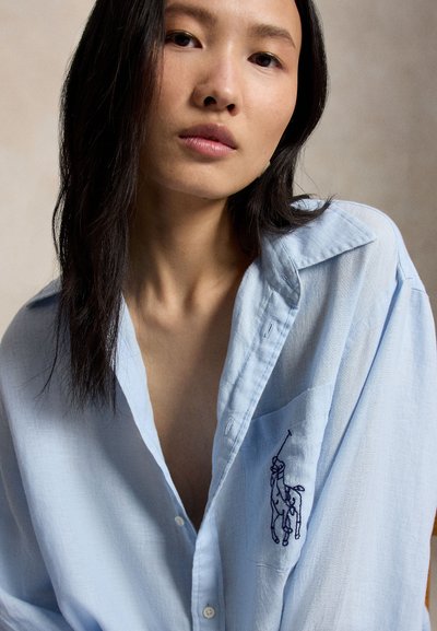 Mladá žena s dlhými tmavými vlasmi, oblečená v svetlomodrej košeli na gombíky s logom pólo hráča na náprsnej vrecku.