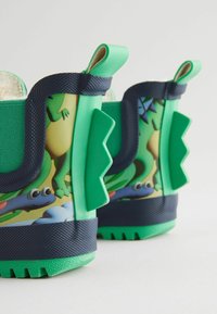 Bottes pour enfants vertes avec un motif de grenouille, en caoutchouc texturé, avec des côtés renforcés et une base bleu marine foncé. Onglets de tirage pour un enfilage facile.
