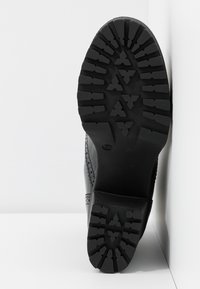 Semelle de chaussure noire avec un motif texturé et robuste, présentant des rainures profondes et un bord plat. Construction solide en caoutchouc avec des détails soignés.
