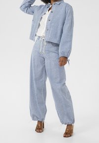 Pruhovaný modro-bílý bavlněný outfit, který zahrnuje zkrácenou bundu se zapínáním na knoflík, kalhoty na stahovací šňůrku a hnědé sandály na pásky.