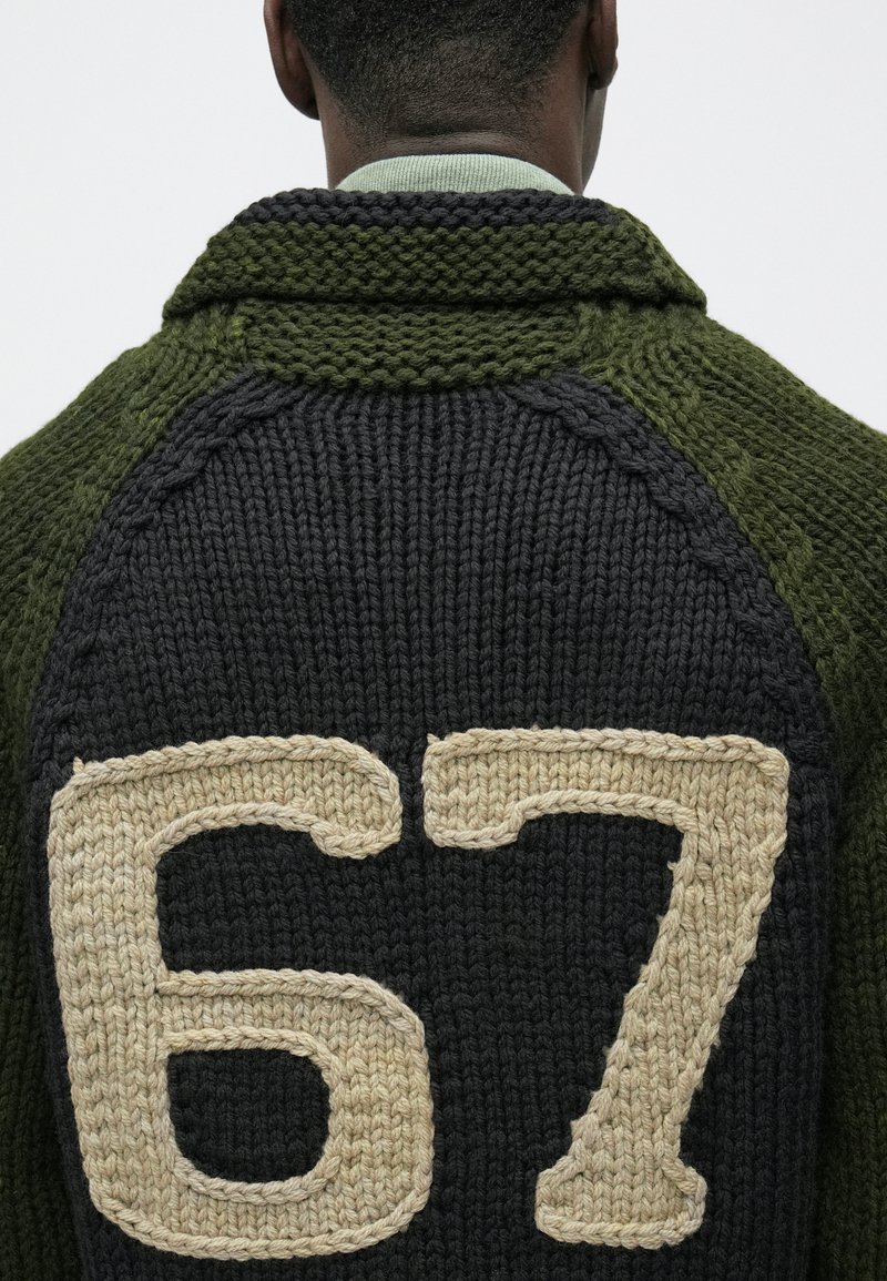 Pull en tricot avec un col et des manches verts, un corps foncé et un grand "67" beige au dos. Tissu en laine texturée.