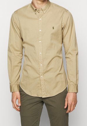 Mand iført en beige skjorte med knapper og lange ærmer med en lille broderet logo på venstre bryst og olivengrønne bukser.