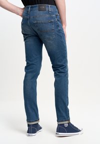 Džíny slim-fit v modré denimové barvě s středním pasem, pěti kapsami a kontrastním šitím. Nosič má na sobě námořnické plátěné tenisky.