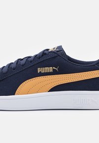 Baskets en suède bleu marine avec une surface texturée, dotées d'une marque dorée et d'une bande orange contrastante sur le côté, semelle blanche avec logo.